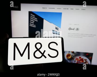 Person mit Smartphone und Logo des britischen Einzelhandelsunternehmens Marks and Spencer Group plc vor der Website. Schwerpunkt auf dem Display des Telefons. Stockfoto