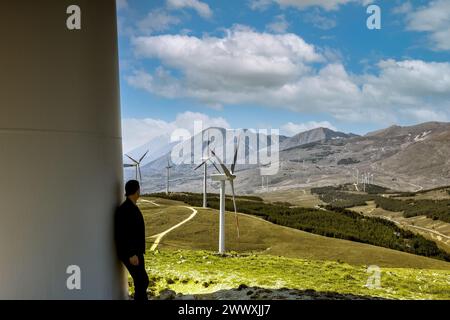 Ein Mann von hinten, der sich am Fuße einer Windkraftanlage stützt, betrachtet die bergige italienische Landschaft mit einem Windpark und in Bewegung befindlichen Turbinen. Ein Wind Stockfoto