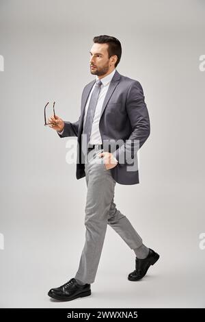 Selbstbewusster Geschäftsmann in formeller Kleidung, der Brille hält und mit Hand in der Tasche auf Grau geht Stockfoto