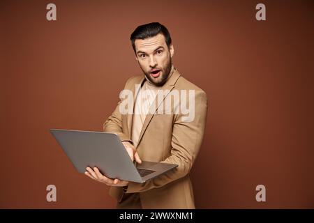 Schockierter und gutaussehender Profi, der sein Notebook benutzt, während er ferngesteuert auf beigefarbenem Hintergrund arbeitet Stockfoto