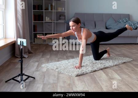 Eine Frau in schwarzer Leggings und weißem Oberteil macht zu Hause Yoga auf dem Teppich und nimmt ein Online-Fitness-Video für ihr Publikum auf, während ein Smartphone steht Stockfoto