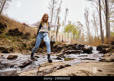 Fröhliche blonde Wanderer in Pullover und Jeans, die über den Waldbach springen und über Wasser springen Stockfoto