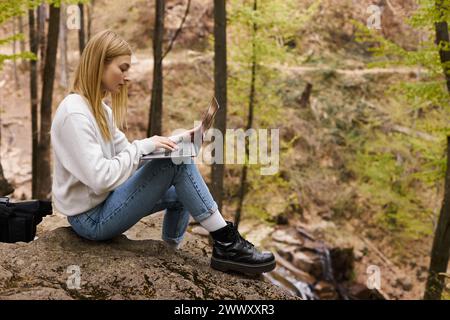 Blonde Abenteurerin, die durch die Wälder wandert, auf Felsen sitzt, mit Laptop und Rucksack Stockfoto