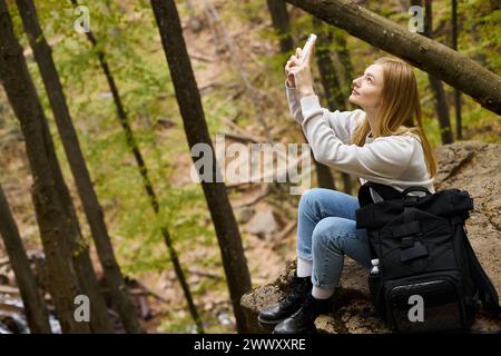 Seitenansicht einer blonden jungen Reisenden, die während der Wanderung Fotos von der Landschaft im Wald macht Stockfoto
