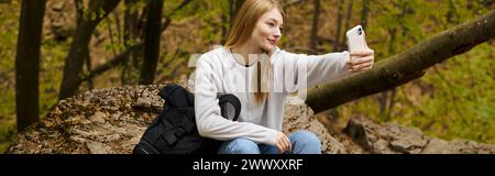 Blonde junge Frau, die Selfie macht, sitzend auf dem Felsen im Wald, während sie wandert, Banner Stockfoto