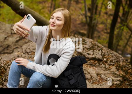 Lächelnde, friedliche, blonde junge Frau, die Selfie macht, während sie sich beim Wandern im Wald entspannt Stockfoto