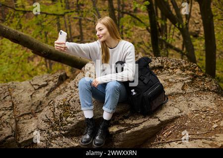Blonde junge Frau, die Selfie macht, sitzend auf einem Felsen im Wald beim Wandern Stockfoto