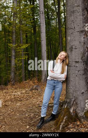 Hochhöhen-Porträt einer blonden Frau in Wanderkleidung im Wald mit geschlossenen Augen auf Baum gelehnt Stockfoto