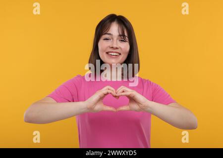 Glückliche Frau, die Herzgeste mit Händen auf orangefarbenem Hintergrund zeigt Stockfoto