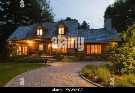 Beleuchtete alte 1800er Jahre rekonstruierte Blockhütte Hausfassade und Pflastersteineinfahrt in der Abenddämmerung, Quebec, Kanada Stockfoto