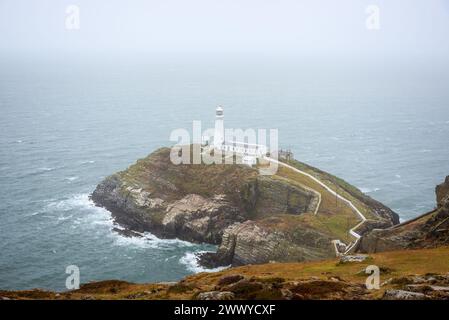 Historischer Leuchtturm auf einer kleinen felsigen Insel vor einer zerklüfteten Küste an einem nebeligen und regnerischen Sommertag. South Stack Lighthouse, Anglesey, Wales, Stockfoto