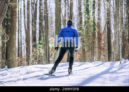 Im Winter gleitet ein einsamer männlicher Skifahrer in einer hellblauen Jacke auf einem schneebedeckten Trail durch den Wald. Ein Athlet läuft Skaten auf Langlaufskiern, Rückansicht. Stockfoto