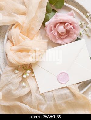 Versiegelter Umschlag in der Nähe von Knoten aus cremefarbenem Tüll und hellrosa Rosen auf den Tellern von oben, Kopierraum. Hochzeit Schreibwaren Mockup. Romantischer Tisch mit Stockfoto