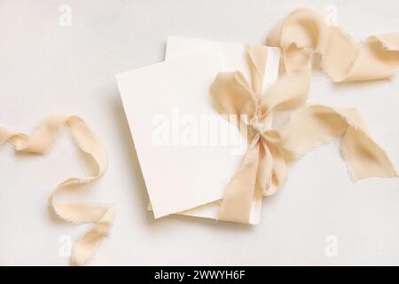 Karten, die mit einem beigefarbenen Seidenband gebunden sind, auf der weißen Tischplatte. Hochzeit Schreibwaren Mockup. Romantische Szene mit vertikalen leeren Karten. Hochzeit, Valentinstag, Stockfoto