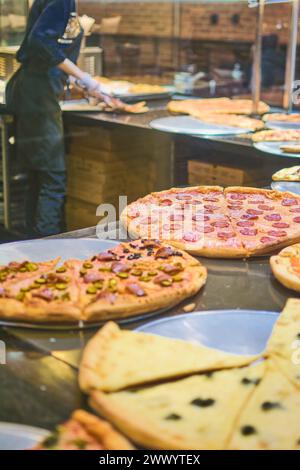 Lebendiges Bild, das eine Vielzahl köstlicher frischer Pizzen zeigt, während ein Koch im Hintergrund in einer beleuchteten Pizzeria mehr zubereitete. Stockfoto