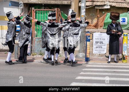 Männer tanzen an einem Quersteg, während ein anderer Mann ein Gebetsbuch liest, während er darauf wartet, während der Purim-Feier zu überqueren. Ultraorthodoxe Juden feiern Purim in Bnei Brak, Israel. Der Feiertag erinnert an die Rettung der Juden im alten Persien durch einen Plan zur Vernichtung. Ein fröhlicher Feiertag, der sowohl von weltlichen als auch von nichtweltlichen Juden gefeiert wird, vor allem indem sie sich in Kostümen kleiden und trinken, so der Talmud, „bis sie nicht unterscheiden können zwischen ‚verflucht ist Haman‘ und ‚gesegnet ist Mordechai‘. Stockfoto