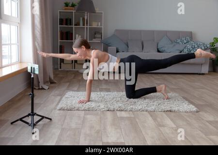 Eine Frau in schwarzer Leggings und weißem Oberteil macht zu Hause Yoga auf dem Teppich und nimmt ein Online-Fitness-Video für ihr Publikum auf, während ein Smartphone steht Stockfoto