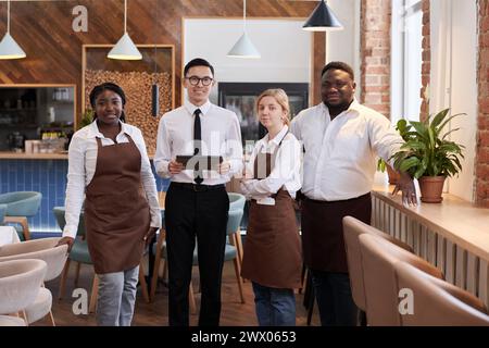Mittellanges Gruppenporträt von jungen, multiethnischen Kellnern, die bei der Arbeit in einem modernen Restaurant vor der Kamera posieren Stockfoto