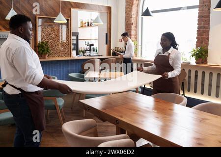 Junger afroamerikanischer Mann und Frau, die in einem modernen Restaurant arbeiten und Tischdecken halten, asiatischer Manager macht Papierkram im Hintergrund Stockfoto