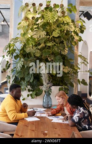 Vertikale Aufnahme kreativer junger afroamerikanischer und kaukasischer Menschen, die während eines Meetings in einem modernen Restaurant mit Kopierraum an einem Geschäftsprojekt arbeiten Stockfoto
