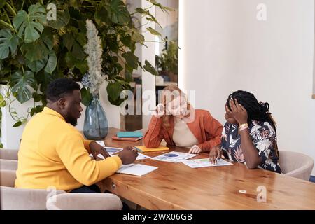Gruppe von drei verschiedenen jungen Geschäftsleuten in stilvoller Freizeitkleidung, die am Tisch im Restaurant sitzen und die Arbeit am Projekt genießen Stockfoto