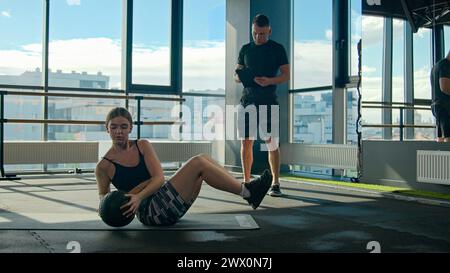 Muskulöse Sportlerin, die Bauchtraining mit Medizinball auf Gym-Boden macht athletisches Frauentraining mit Mann, persönlichem Sporttrainer, Fitnessmädchen Stockfoto