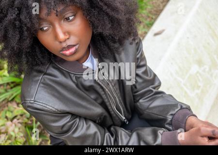 Ein Porträt einer kontemplativen afroamerikanischen Frau, die draußen sitzt, mit ihren ausdrucksstarken Augen und der natürlichen Locken ihres Haares vor einem weichen, natürlichen Hintergrund. Sie trägt eine Lederjacke, was eine Mischung aus Nachdenklichkeit und Stil andeutet. Nachdenkliche afroamerikanische Frau mit natürlichem Haar im Freien. Hochwertige Fotos Stockfoto