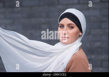 Porträt einer jungen blauäugigen Frau in einem Hijab vor einer grauen Ziegelwand. Weißer Schal flattert im Wind. Stockfoto