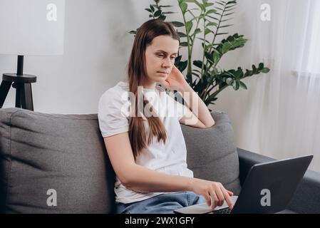 Fokussierte freischaffende Mädchenarbeit am Laptop auf gemütlicher Couch in modernem Wohnzimmer, sieht durchdachte Such-Ideen aus, korrespondiert mit dem Kunden, löst Bus Stockfoto