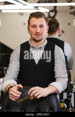 Ein Friseur in Hemd und Weste steht neben einem Friseurstuhl, der Werkzeuge für Haarschnitte hält. Hände des Friseurs halten eine Schere und einen Kamm gegen den Stockfoto