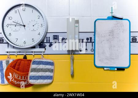 An der Wand hängt ein Whiteboard mit blauem Rand. Es ist leer und weist leichte Gebrauchsspuren auf. Weiße Uhr mit schwarzen Zahlen und Zeigern hängt an einem gelben Stockfoto