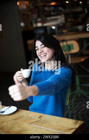 Eine fröhliche Frau in einem blauen Pullover genießt eine Tasse Kaffee in einem Café im Innenbereich und gibt mit einem hellen, fesselnden Lächeln die Daumen hoch Stockfoto