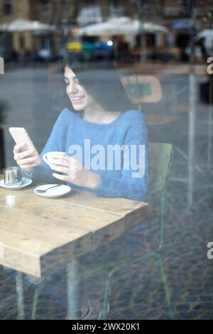Eine junge Frau in einem blauen Pullover fokussiert sich in einem Café auf ihr Handy, mit einer Kaffeetasse und Untertasse vor ihr, durch ein Fenster gesehen Stockfoto