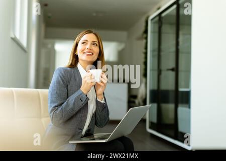 Die professionelle Profi schlürft ihren Kaffee, während sie sich mit ihrem Laptop beschäftigt, während sie sich bei weichem Tageslicht sonnt. Stockfoto