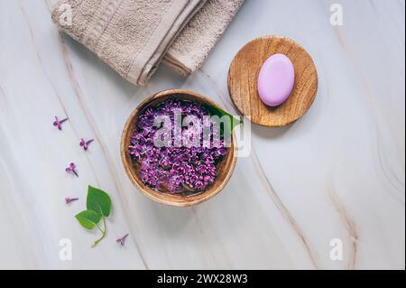 Spa- und Wellness-Komposition mit duftendem Fliederblütenwasser in einer Holzschale, Frottierhandtüchern und handgemachter Seife auf Marmorhintergrund. Aromatherapie, oben Stockfoto