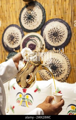 Ein arabischer Mann in weißem Kandura, der arabischen Kaffee aus einer traditionellen Teekanne, dem Wasserkocher Dalla in Katar, in eine Einwegbecher gießt. Stockfoto