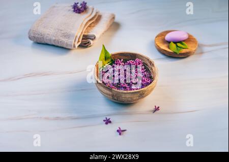 Spa- und Wellness-Komposition mit duftendem Fliederblütenwasser in einer Holzschale, Frottierhandtüchern und handgemachter Seife auf Marmorhintergrund. Stockfoto
