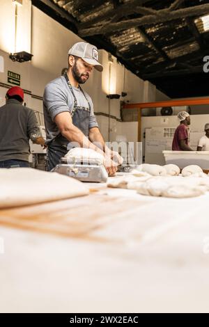 Junger erwachsener Bäcker, der Teige zubereitete, um Brot auf dem Bäckereitisch zu machen, die Teige wog, Bäckereischule. Stockfoto