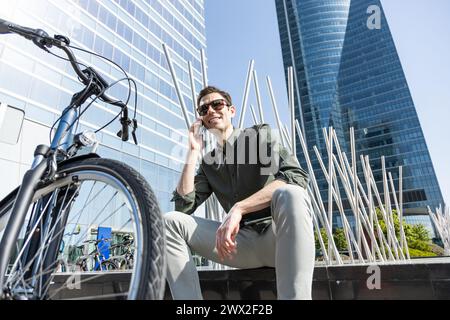 Ein lächelnder junger Geschäftsmann fährt E-Bike auf dem Weg zum Turm, in dem sich sein Büro befindet, und führt ein Gespräch mit Ihrem Smartphone Stockfoto