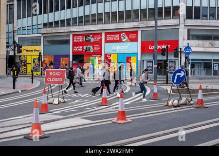 Menschen, die während der Baustelle zwischen Verkehrskegeln um eine Kreuzung laufen, während der Modernisierung des öffentlichen Nahverkehrssystems der Midland Metro im Stadtzentrum in der Nähe der Corporation Street am 21. März 2024 in Birmingham, Großbritannien. Die ursprünglichen Gleise werden hochgezogen und umgebaut, während eine neue Strecke im Bau ist und später im Jahr eröffnet werden soll. Die Midland Metro ist eine Straßenbahnlinie im County West Midlands, England, die zwischen Birmingham und Wolverhampton über die Städte West Bromwich und Wednesday verkehrt. Die Strecke verkehrt auf den Straßen in Stockfoto