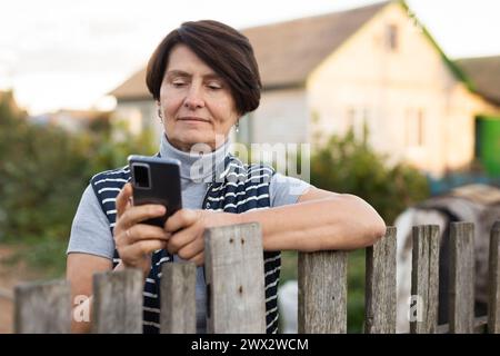 Reife Frau, die entspannt neben dem Zaun steht und am Telefon im Garten lächelt Stockfoto