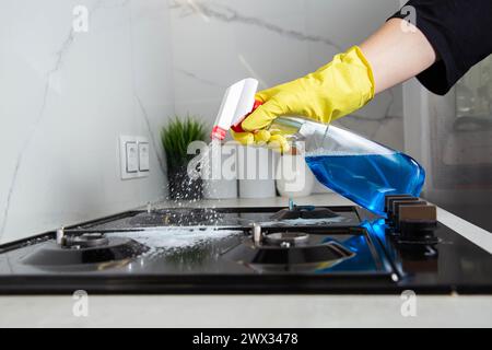 Die Hand eines Mädchens mit einem Reinigungsspray sprüht auf einen Gasherd. Modernes fettfreies, antistatisches Reinigungsmittel. Hintergrund: Weiße Porzellanfliesen Stockfoto