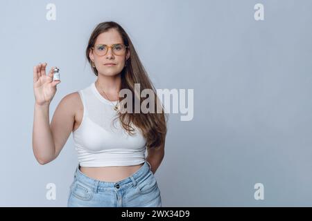Junge lateinfrau mit Brille und langen Haaren, Kosmetikerin Arzt stehend posiert Ampulle für botox-Behandlung, Studioaufnahme mit weißem Hintergrund Stockfoto