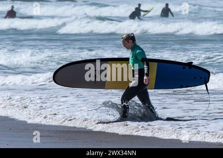 Eine reife Surferin, die ihr Surfbrett trägt und aus dem Meer in Fistral in Newquay in Cornwall in Großbritannien spaziert. Stockfoto