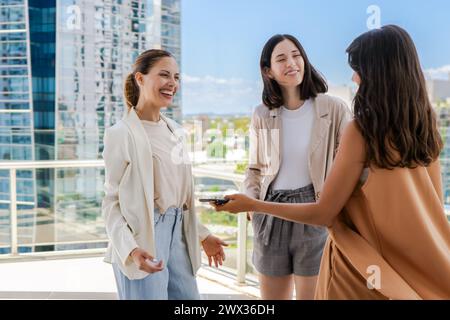 Drei Frauen stehen auf einem Dach, lächeln und reden miteinander. Einer von ihnen hält ein Handy Stockfoto