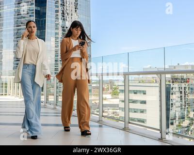 Zwei Frauen laufen auf einem Glassteg. Einer von ihnen spricht am Telefon, der andere sendet eine Nachricht. Die Szene spielt in einer Stadt mit Tal Stockfoto