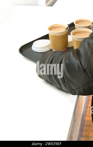 Ein in einer schwarzen Jacke gekleideter Arm hält ein Tablett mit vier Tassen Kaffee zum Mitnehmen, was einen schnellen und effizienten Service ermöglicht. Stockfoto