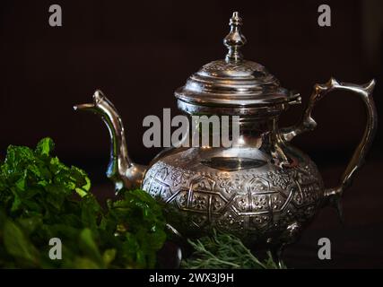 Eine silberne Teekanne und ein Haufen frischer grüner Minze im Vordergrund. Traditioneller marokkanischer Tee als Symbol der Gastfreundschaft der marokkanischen Menschen. Selektiv Stockfoto