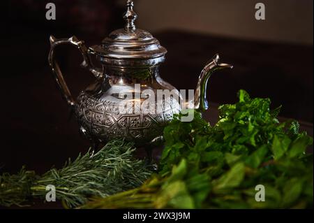 Eine silberne Teekanne und ein Haufen frischer grüner Minze im Vordergrund. Traditioneller marokkanischer Tee als Symbol der Gastfreundschaft der marokkanischen Menschen. Selektiv Stockfoto