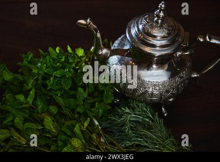 Eine silberne Teekanne und ein Haufen frischer grüner Minze im Vordergrund. Traditioneller marokkanischer Tee als Symbol der Gastfreundschaft der marokkanischen Menschen. Selektiv Stockfoto